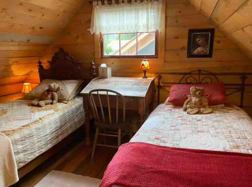 Cozy attic bedroom with two beds, a wooden desk, and teddy bears, featuring rustic wooden walls and warm lighting.